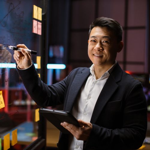 Asian businessman planning project on glass board in office Confident male ceo manager, writing tasks or new startup ideas on stickers on glass wall, while checking project progress using tablet pc. Asian businessman planning project on board in office